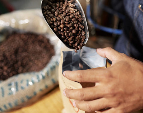 Hands pour roasted coffee beans from a metal scoop into an open bag. A burlap sack filled with more beans is visible in the background. Warm, artisanal setting.