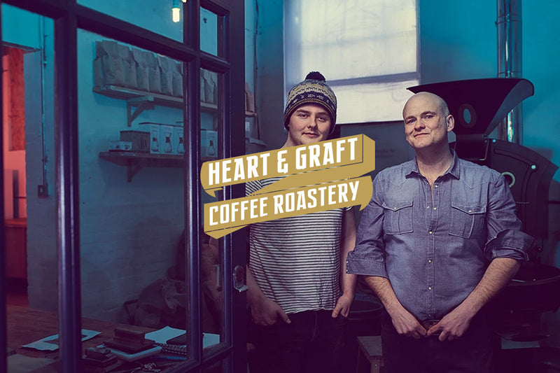 Two people stand in a cosy coffee roastery with a warm glow. One wears a striped shirt and beanie, the other a denim shirt, both looking content. Text reads "Heart & Graft Coffee Roastery."